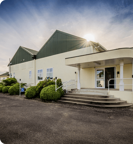 A cream-coloured building with green gabled roofs, front steps with railings, neatly trimmed shrubs, and the sun shining from behind the roofline.