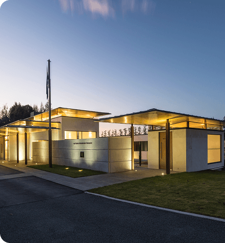 A modern, low-profile building with warm exterior lighting at dusk, featuring concrete walls, wooden beams, and a flagpole at the entrance.