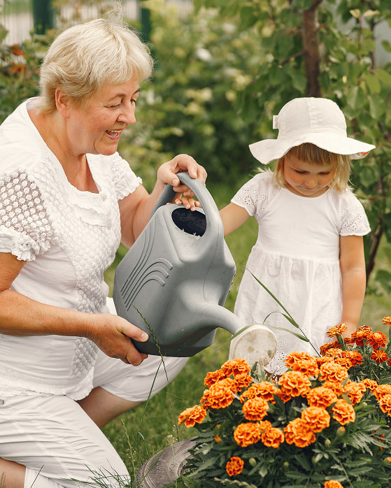 An older woman and a young girl dressed in white watering vibrant orange flowers together in a garden.