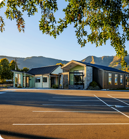 A contemporary building in a wide parking area with mountains and trees in the background, lit by early morning or late afternoon sunlight.